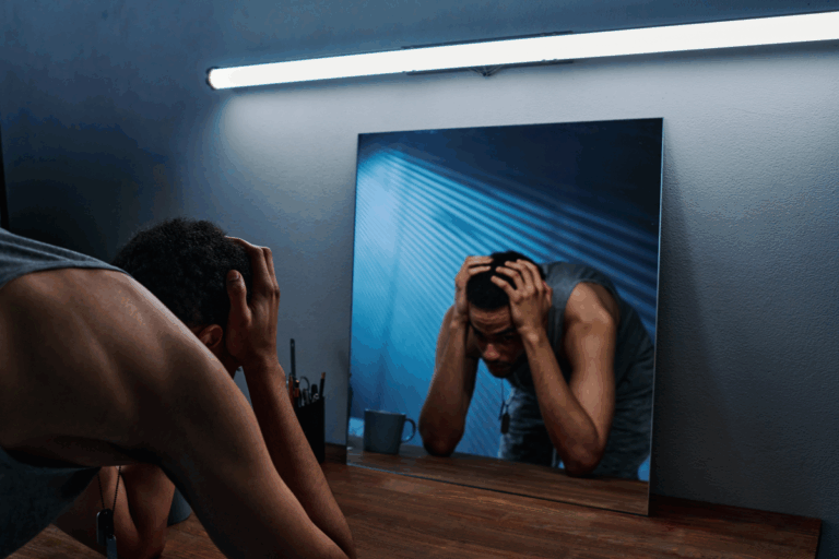 Man leaning over a table with his head in his hands, reflected in a mirror, illustrating emotional distress linked to complex PTSD (CPTSD)