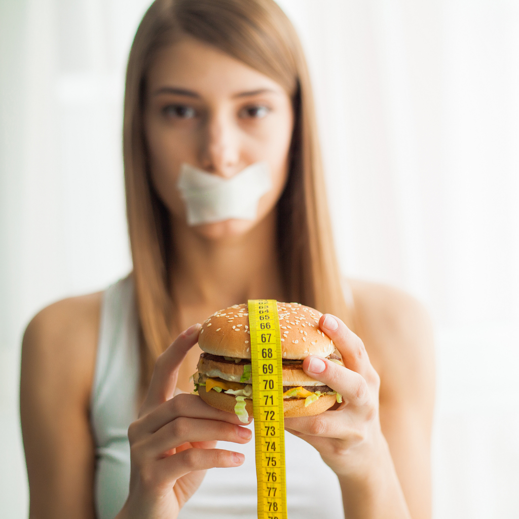 Social Media's Influence on Body Image and Eating Disorders 2 Young woman with tape over her mouth holding a burger wrapped in a measuring tape, symbolizing representing unhealthy dieting and pressure to lose weight.