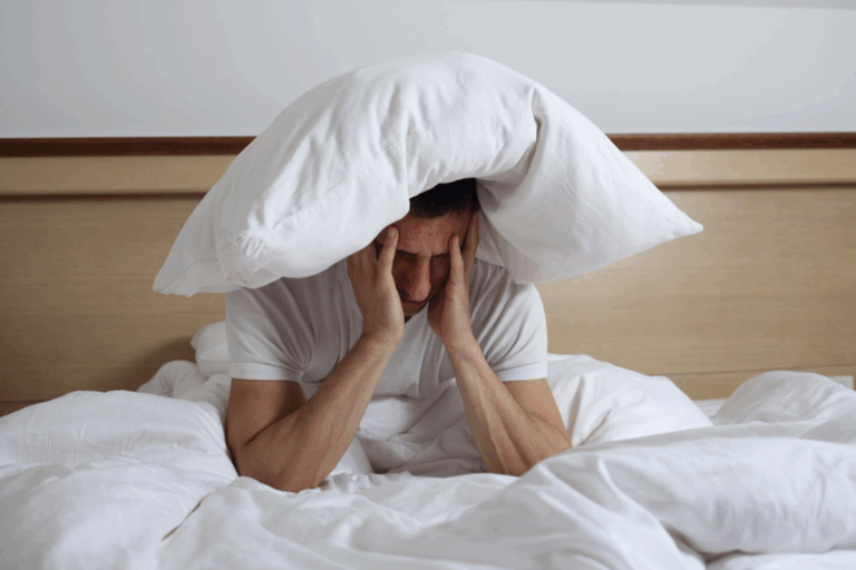 Man struggling with insomnia holding his head under a pillow in bed, showing frustration and lack of sleep.