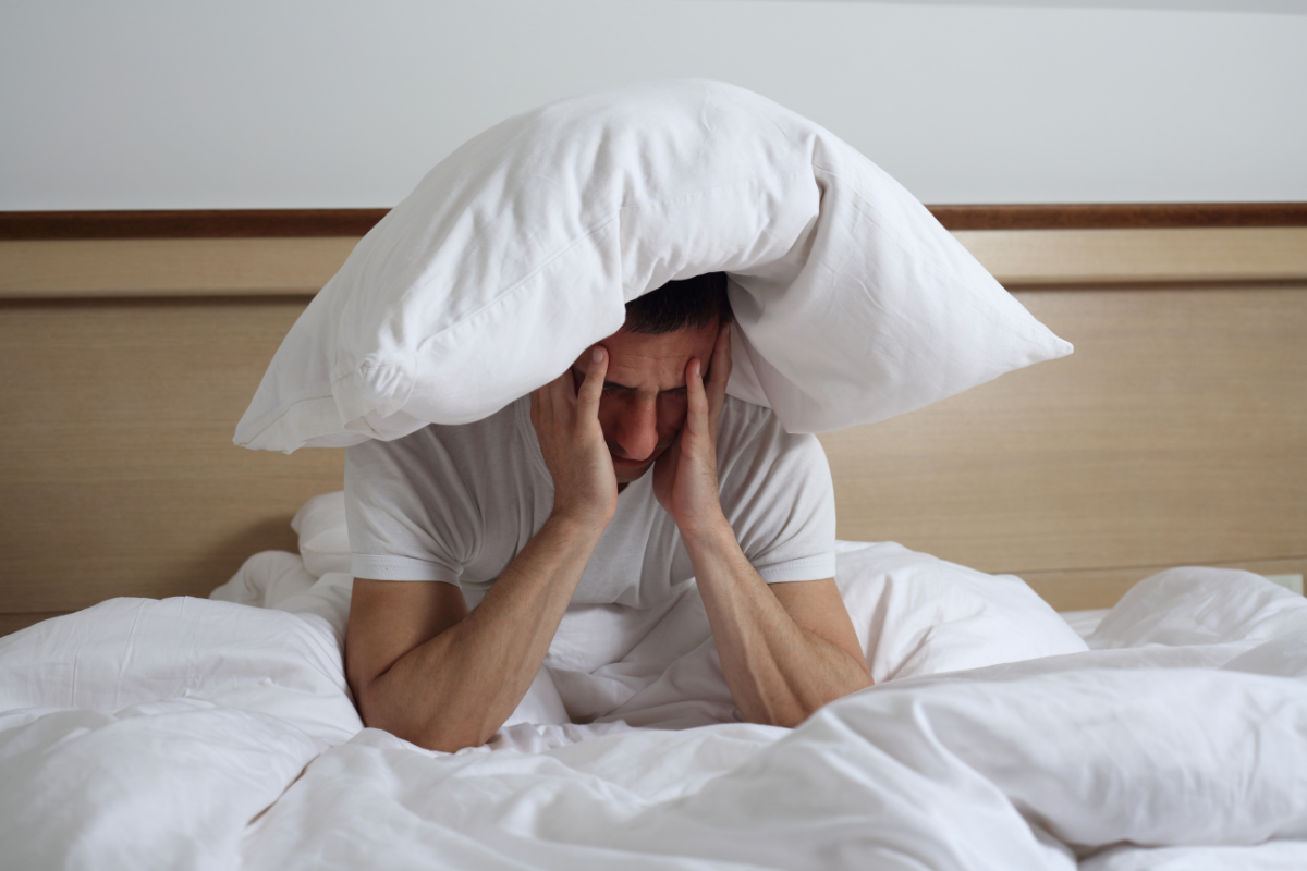 Man struggling with insomnia holding his head under a pillow in bed, showing frustration and lack of sleep.