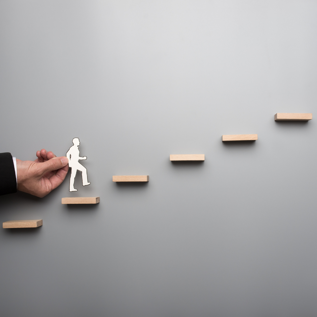 Hand moving paper cutout of person up wooden steps, showing step-by-step progress.