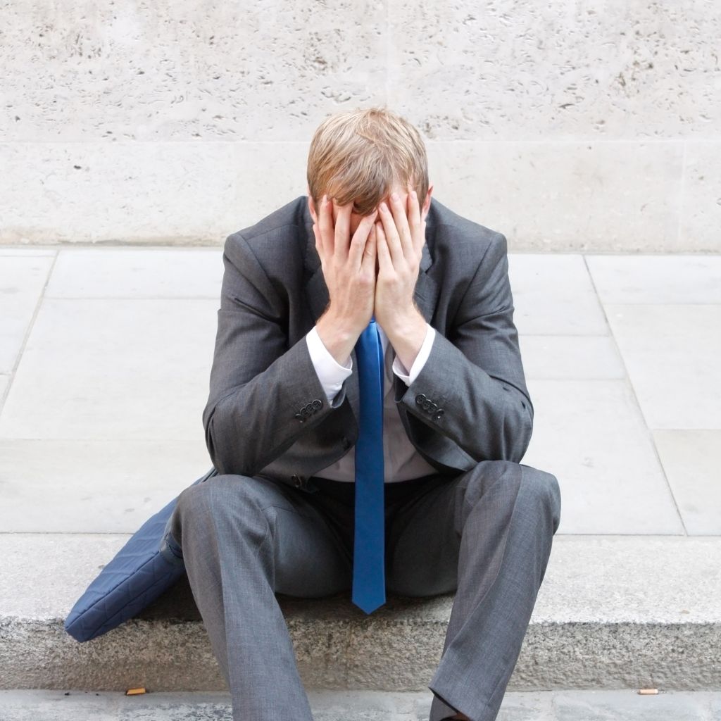 Imposter Syndrome and Mental Health: The Hidden Cost of Self-Doubt 1 Man in a suit sitting on the ground with his head in his hands, showing signs of stress and self-doubt often linked to imposter syndrome.