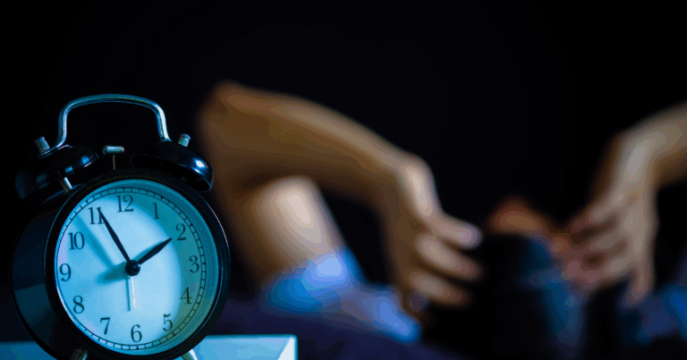 Alarm clock on a bedside table with a person struggling to fall asleep in the background.