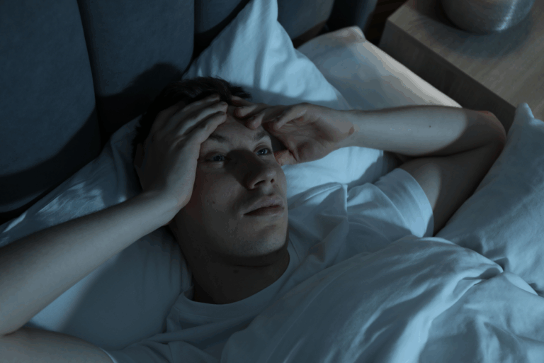 A man lying awake in bed at night, holding his head as anxiety and insomnia keep him from sleeping.