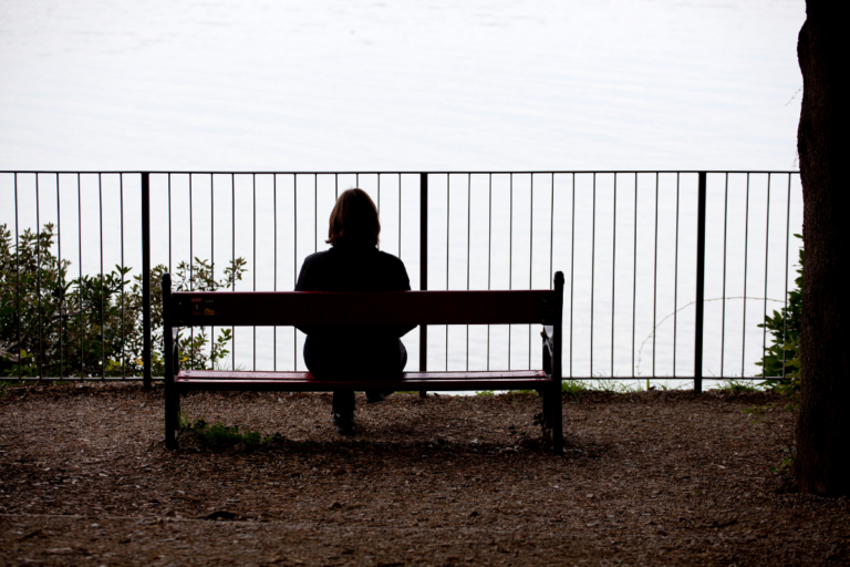 A person sitting alone on a bench, looking out over the water, reflecting emotional withdrawal and early signs of depression relapse.