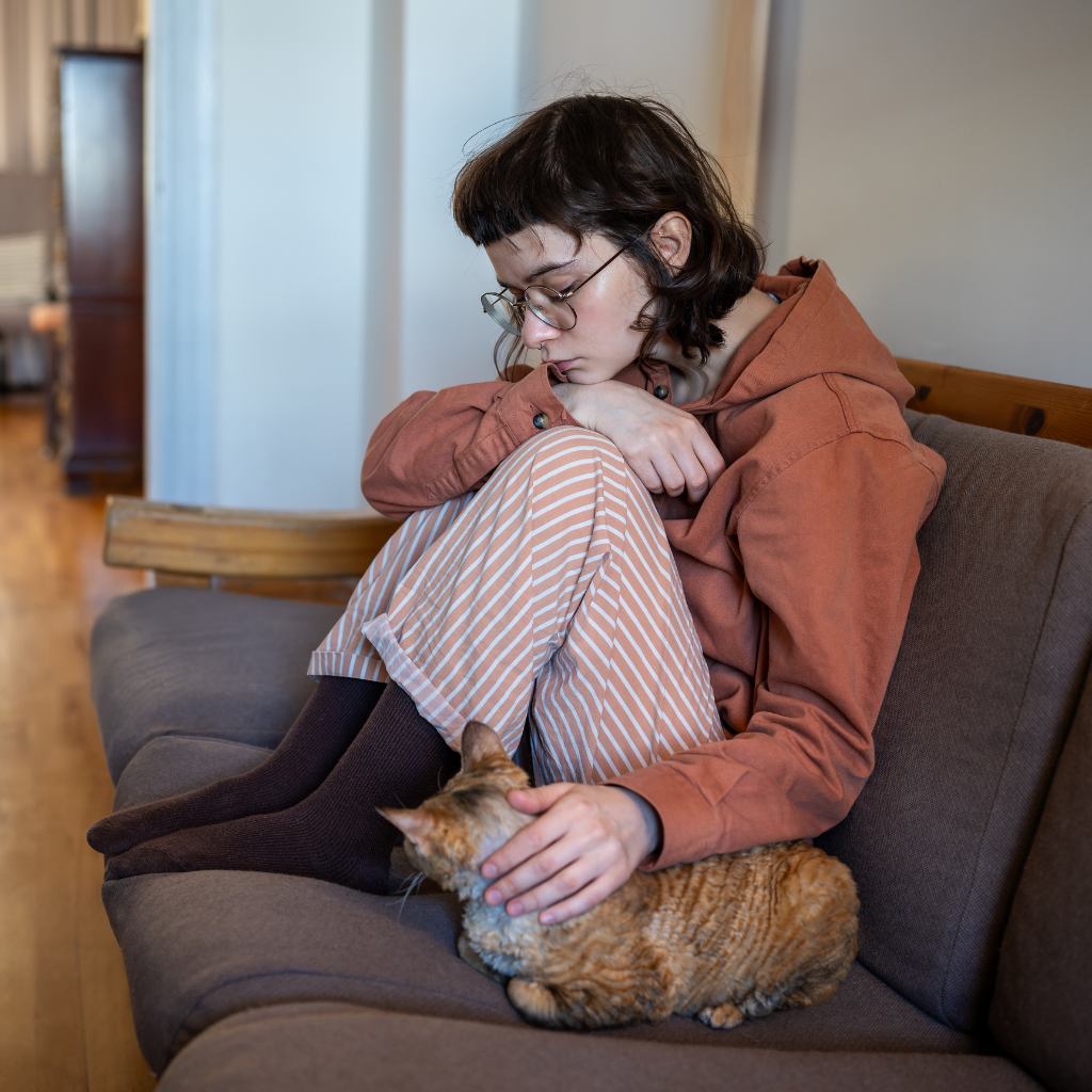 A person sitting curled up on a couch, gently petting a cat, appearing withdrawn and low in mood.