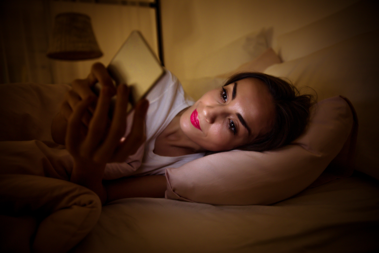 Woman using her phone in bed at night despite feeling exhausted.