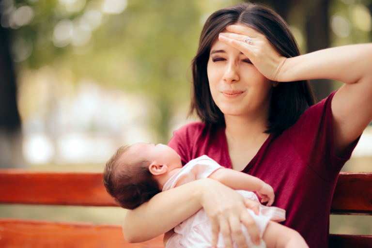 New mother holding baby outdoors with hand on forehead, reflecting postpartum anxiety symptoms after birth.