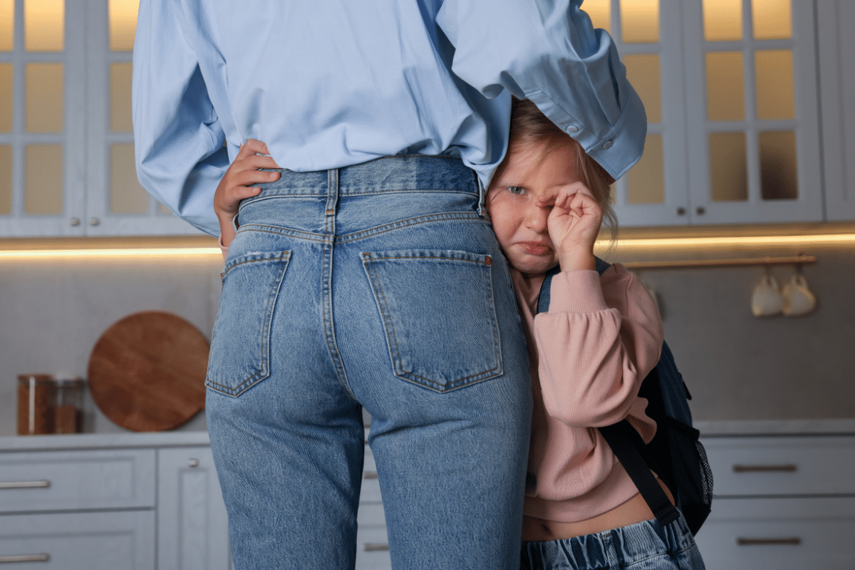 Young child with a backpack hiding behind a parent, showing how anxiety in young children can look like clinginess before school.