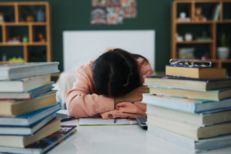 Teen student resting her head on a desk between stacks of books, showing exhaustion from academic pressure and schoolwork.