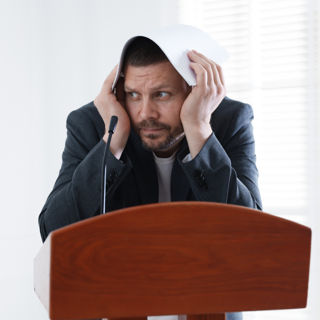 Man at a podium covering his head with papers, appearing overwhelmed and fearful during a work presentation.