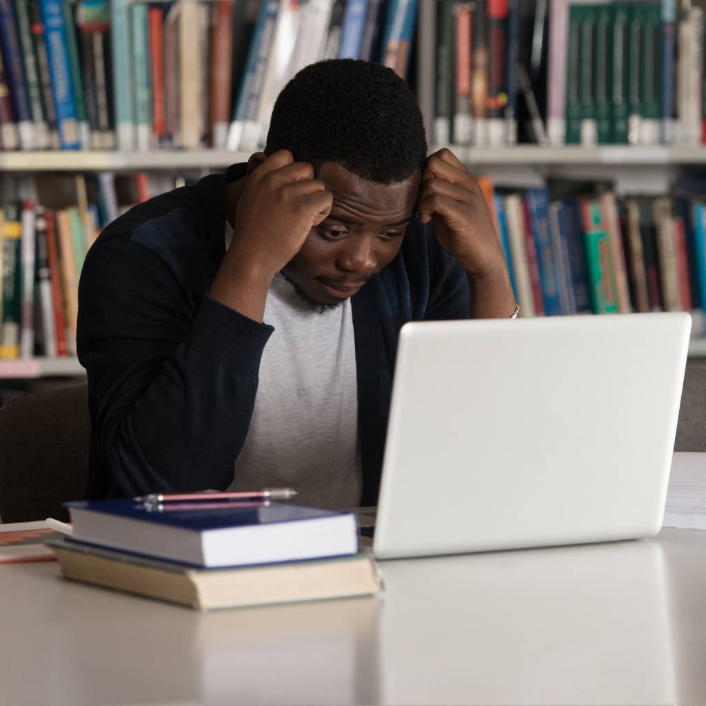 Student sitting in a library with a laptop and books, appearing stressed and overwhelmed while studying.