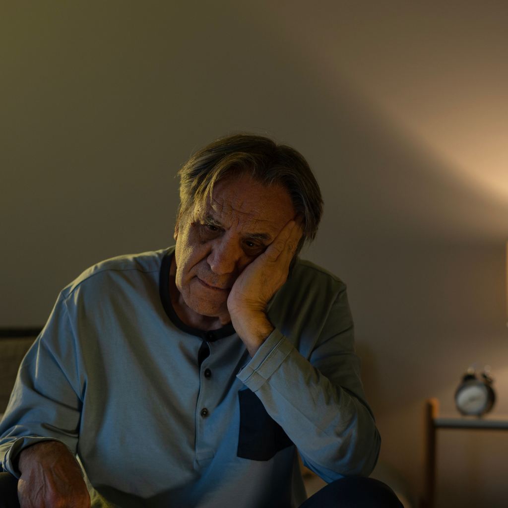 Older man sitting awake at night looking tired and restless, reflecting common sleep difficulties in later life.